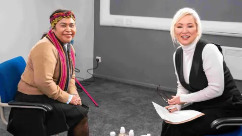 FSWC Two woman are sitting in chairs opposite each other. They are both looking into a camera and smiling. The woman on the left is wearing a red and yellow head scarf, pink scarf, brown cardigan and black skirt. The woman on the right has tied back bright blonde hair. She is wearing a white jumper, black top and black trousers.