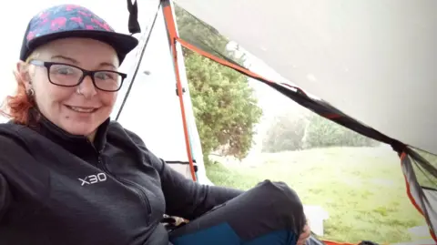 Becky Harrison sitting inside a tent while wild camping. Ms Harrison is smiling at the camera. The tent door is open and is showing a field and trees.