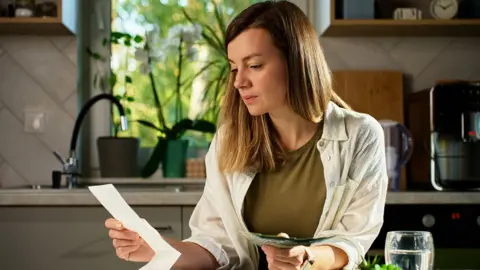 A woman sitting at a table and looking at houshold bills in her kitchen . In the background is a window showing green plants and trees