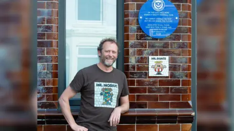 Mr Mosaic Will Rosie wearing a t-shirt which says "Mr Mosaic" he is leaning on the wall where there is a blue plaque which shows the Portsmouth Football Club crest above text which reads "on this site once more stands a mighty football club. We cannot change the past but we can shape the future. Dedicated to all those fans who took a stand and refused to allow Portsmouth FC to die" underneath the plaque is a white mosaic with a mr man character dressed in a southampton fc kit, it says "mr saints" at the top