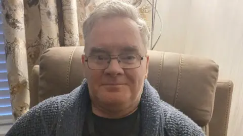 Gerry Rea is seen wearing a blue knitted cardigan and black t-shirt. 
He has black glasses and is sitting in a tanned chair near a window