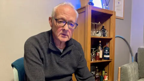 A man with silver hair wearing glasses and a brown polo top.