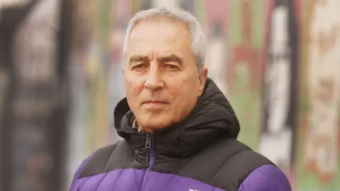 Sinn Féin An older man with short white hair. He is staring towards the camera and is wearing a black and purple puffy hooded jacket zipped up to his chin. 
