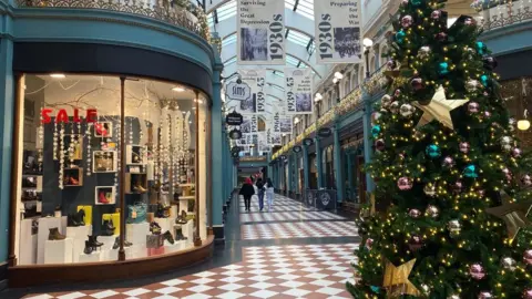 The inside of a shopping arcade has orange and white checkered tiles on the floor with a Christmas tree on the middle of the floor. There are shop windows full of shoes and Christmas gifts 