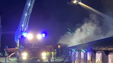 ECFRS A fire engine has all of its lights on and an aerial ladder extended from its rear. Smoke is on one side of the picture and blue lights are on the fire engine.