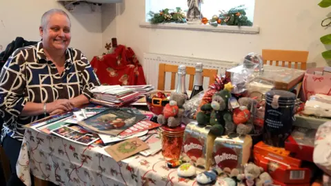 Photograph of Clare Jones sat at a table filled with Christmas cards and gifts. She sits on the far end of the table on the left and smiles down at the cards. She has short grey hair and wears a brown, white and navy shirt. On the table are an array of stuffed animals, chocolates and bottles of prosecco. Several cards are stacked in front of Clare. 