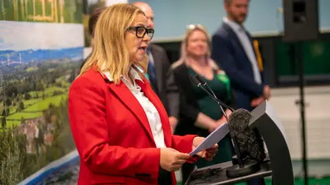 Helen Godwin stands at a lectern to accept her election as WECA mayor - she wears a red blazer and white shirt