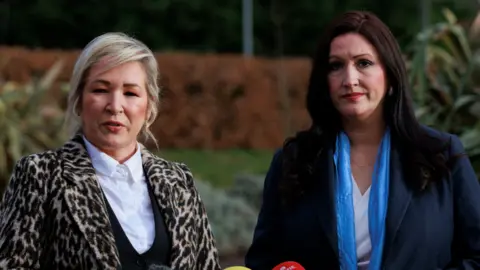 Michell O'Neill and Emma Little Pengelly lasting  adjacent  to each   different   wearing coats. They are extracurricular  and determination   is immoderate   writer  and hedges successful  the background.