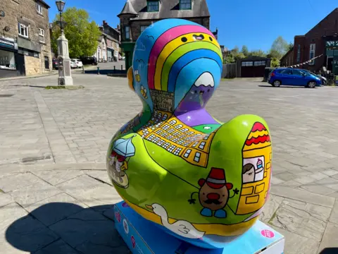 A giant duck sculpture sits in a cobbled square with old buildings and an old street lamp. The duck includes bright cartoons of a mill in Belper, a river, a rainbow, a swan, a swan, a Mr Potato Head and a mountain with a smiley face. It sits on a blue plinth with information printed on it.