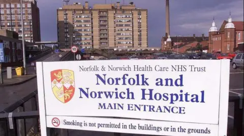 PA Media A picture from 2000 shows a white sign in the foreground which states "Norfolk and Norwich Hospital main entrance" alongside a sign stating that smoking is not permitted. Large concrete hospital buildings and a car park are seen in the background.