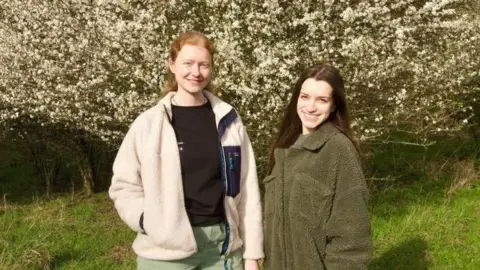Two women stood in front of a blossom tree. The woman on the left has red hair with a cream jacket and black t-shirt with light green trousers. The woman on the right has long brown hair and is wearing a green full-length coat