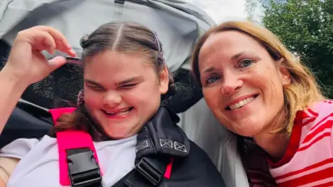 Liz Chaplin Liz Chaplin leans in towards Ava, who is sitting down. Both are smiling in this outdoor scene. Liz is wearing a pink and red-striped jumper, while Ava is wearing a lilac T-shirt.