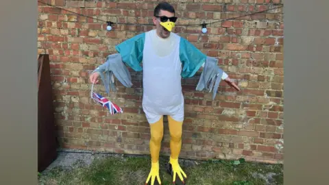 Sam from Rochester dressed as a seagull. Sam is wearing a white overshirt, yellow tights, yellow claw shoes and a beak. He is holding a Union Jack flag.