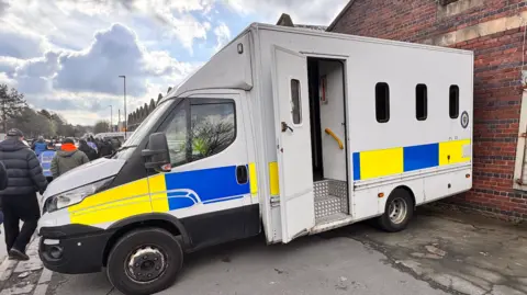 UNITE A police van parked up against a brick building. People can be seen walking on the street alongside it