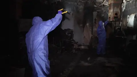 Two people in blue protective overalls and white masks looking through the remains of the burnt out ward