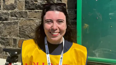 Keely is smiling. She has brown hair. She's wearing a yellow bib that says volunteer. She's outside.