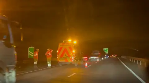 A highway maintenance vehicle is stopped on a dual carriageway. Traffic cones have been placed on the road and two men in high vis are standing at the side of the road talking to each other. 