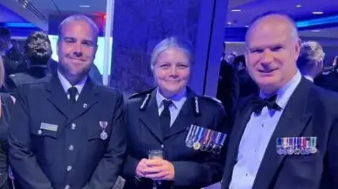 Avon and Somerset Police Justin Robbie, Sarah Crew and Mark Shelford at an awards ceremony, smiling. The three are wearing different numbers of medals and Sarah is holding a champagne flute.