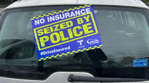 Silver car with a blue yellow and white sticker covering the rear window saying it's been seized by police for having no insurance.  