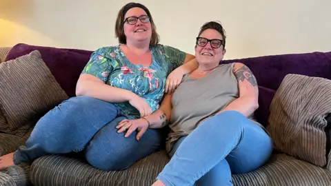 A woman with a black headband and glasses in a blue and pink top and blue jeans sits on a sofa leaning on a woman with short dark hair and glasses, wearing a sleeveless grey vest top and blue jeans. She has tattoos on her upper arm and inner arm.