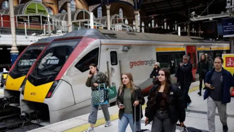 Passengers disembark from a Greater Anglia Class 745 InterCity train at London Liverpool Street station. The train has a yellow, red and grey livery and is a bit dirty looking. Some of the passengers are carrying bags or pulling suitcases on wheels. One is on a mobile phone call.