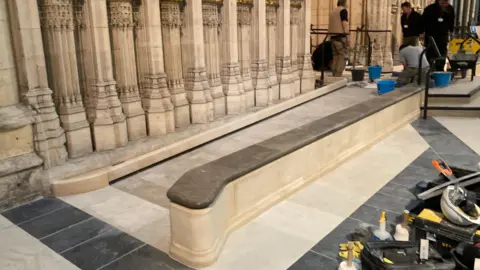 A long, carved stone ramp runs alongside an ornate wall with detailed vertical columns and decorative patterns. Several workers are installing or repairing the flooring, using buckets, tools, and equipment spread across the area. The setting appears to be a historic or grand indoor space, possibly undergoing restoration or maintenance.