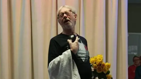 Supplied Tim Strugnell performing on stage. He is holding a bouquet of yellow flowers and wearing a black top with a towel draped over his arm like a waiter.