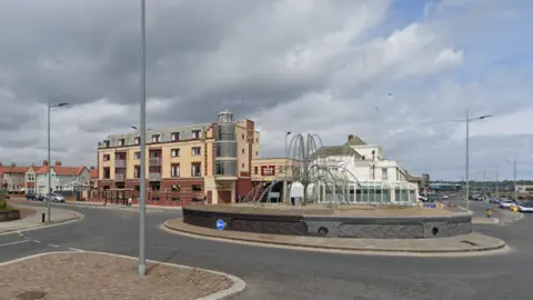 A roundabout on the A183 Whitburn Road. There is a block of apartments in the background and building styles commonly seen at the seaside. The centre of the roundabout has a steel sculpture that resembles the jets of a fountain.