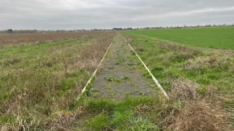 The community path near North Angle Solar Farm. It is surrounded by fields and ends abruptly. It has weeds growing through it