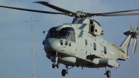 RNAS Culdrose A grey Merlin helicopter in flight with a navigation tower behind it on a blue sky day.