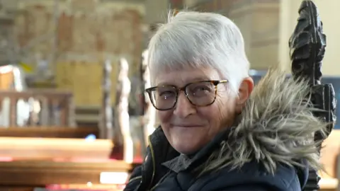 Faith Marsden is sitting on a pew in the church and is smiling. She has grey hair and is wearing a puffer coat with faux fur trim round the hood.