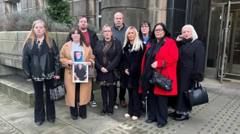Nine people are standing outside St Andrew's House. One is wearing a bright red coat.