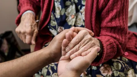 Getty Images Hands holding the hands of an elderly woman. She is wearing a red cardigan and floral dress.