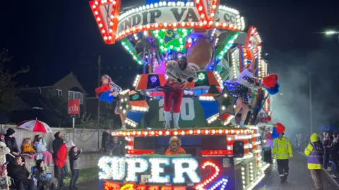 A carnival cart is lit up and coming down a road in Glastonbury. There are spectators at the side of the road.