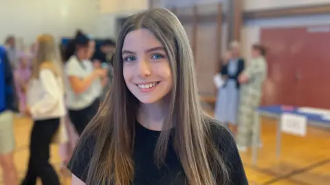 Wiktoria is smiling at the camera. She has long brown hair and is wearing a black top.