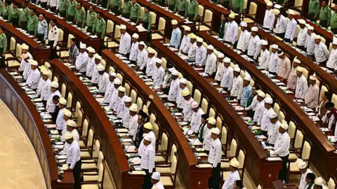 AFP via Getty Images Rows of men in military uniform and white shirts stand behind long tables in Myanmar's parliament.