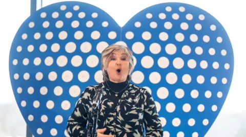 PA Media Lesley Garrett, a woman with a black and white patterned jacket, is stood in front of a blue heart shape. She is singing into a microphone in front of her.