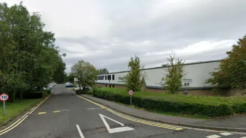 A factory building sits on the right hand side of the road lined with bushes and trees. 