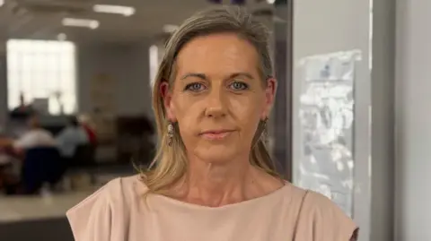 A blonde woman looking at the camera. She is wearing a beige top.