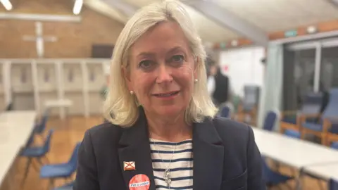 Rhys Thomas/ BBC News Helen Miles has shoulder length blonde hair and is wearing a navy jacket and navy and white striped top. she is standing in a hall with trestle tables and chairs behind her.