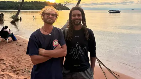 Contributed Two bearded men stand on a beach at sunset together with the water behind them. The man on the left wears a navy T-shirt and has blonde hair and glasses. The other on the right has long dark dreadlocks and wears a black T-shirt with grey shorts. There is a small boat moored offshore and a wooded area on a far shore behind them, across an expanse of water.