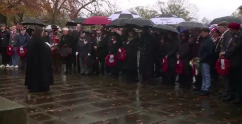 A large group of people gathered on a paved area outside. Many are holding black umbrellas, while others have red poppy wreaths in their hands. Several individuals wear formal attire, including dark coats and military uniforms with medals.