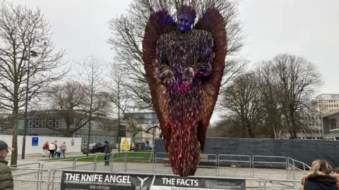 The Knife Angel sculpture in Plymouth. People are stood next to the artwork made from 100,000 blades. Metal fencing has been placed around the sculpture.