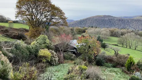 House in the middle of countryside surrounded by trees with autumn leaves and fields. Hills are visible in the background.