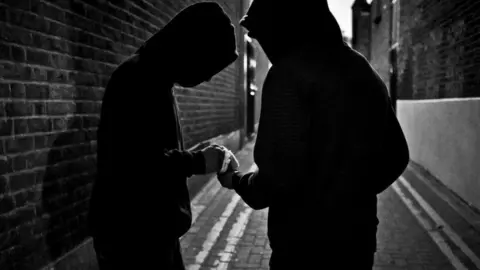 Two boys in shadow in a alley with brick walls and cobbled stoned streets. They are both wearing hoodies and are passing something to each other. The image is portraying a back alley drug deal. 