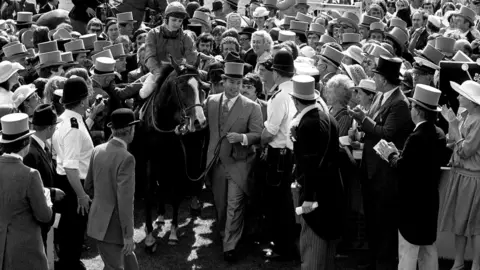 PA It is a black and white image of a crowd of dozens of people surrounding a horse with a white stripe on the front of its face. A jockey is sitting on the horse and a man wearing a top hat and morning suit is leading the horse through the crowd.