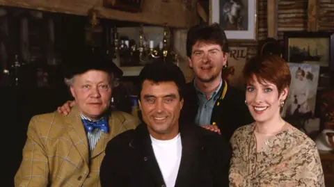A group photo of Chris Jury, Dudley Sutton, Ian McShane and Phyllis Logan. They are standing in a room which appears to be filled with antiques and old items and trinkets. They are all smiling and looking directly into the camera. 
