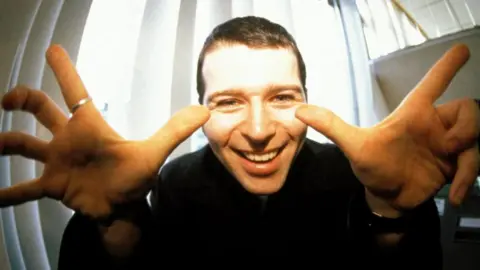 Mick Hutson/ Getty Images Shows a man smiling with his fingers pointing upwards. He is wearing a black top.