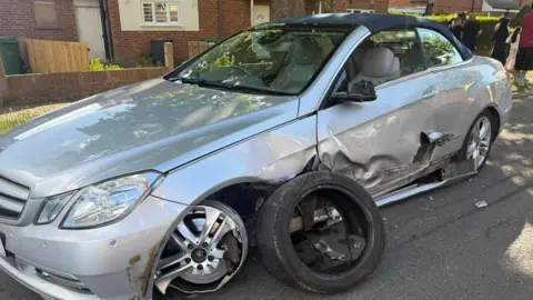 Ishfaq Hussain A side view of a silver car after a crash at the side of a road. Its front wheel hub is buckled and the front tyre has come off and is resting against the side of the car. Its wing mirror is smashed and there is a deep gouge in the side of its passenger side door. Beyond it is a grass verge and beyond that rows of houses. 
