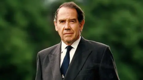 Sir Terence walks towards the camera looking at the lens, with one eyebrow slightly raised. We can just see him from the chest upwards. He wears a grey suit jacket, white shirt and blue tie. He has short dark hair and is clean shaven.